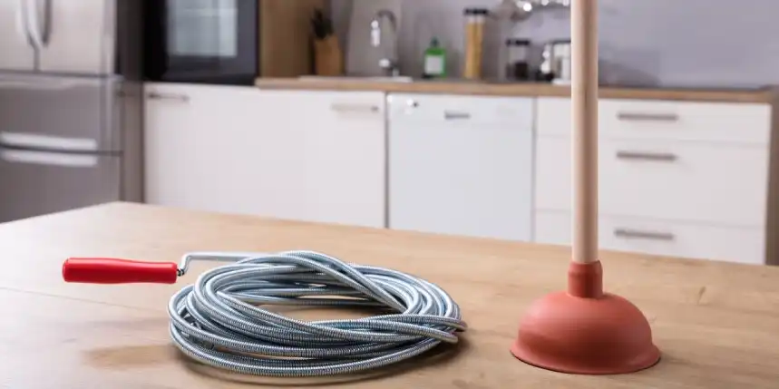 A coiled, metal drain snake and a plunger sitting on a counter before being used to unclog a drain in Calgary, AB.