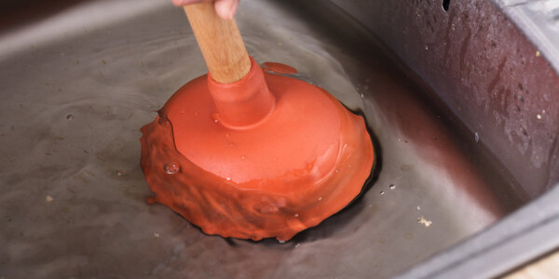 A plunger covering the drain opening of a metal sink filled with water as it’s being used to unclog the sink.
