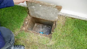 A plumber's hand holding open a home's sewer cover to expose sewage water clogging the pipes.