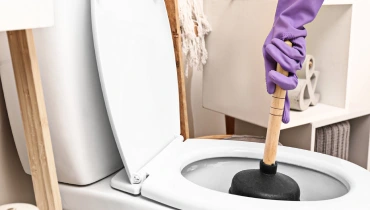 A plumber wearing rubber gloves and holding a plunger in the bowl of a toilet as they clear a toilet clog.