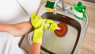 A close-up image of someone wearing rubber gloves and using a plunger to clear a clogged sink.