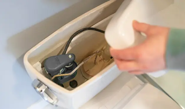 A person removing the lid of their toilet tank to investigate the interior of the tank, revealing the float and flapper valve in the tank below. 