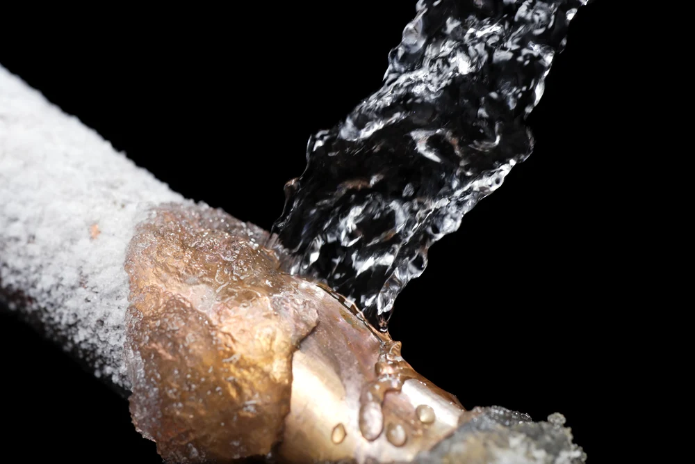 A copper  pipe covered in frost with water shooting out of a burst on the side that must be fixed with frozen pipe repair services.