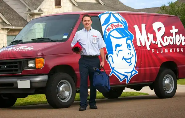 Mr. Rooter plumber standing in front of van parked on street, holding mat and utility bag.