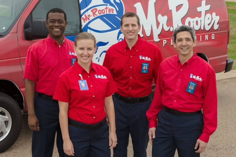 A team of smiling plumbers from Mr. Rooter Plumbing standing in front of a branded, Mr. Rooter Plumbing van before providing frozen pipe repair services.