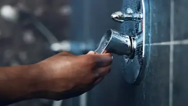 A close-up of a person's hand adjusting a chrome shower faucet handle on a tiled bathroom wall.