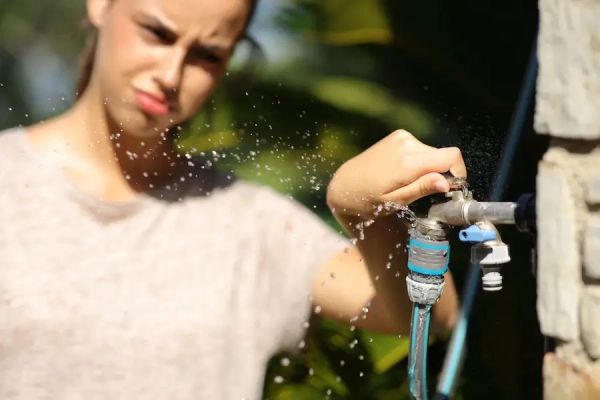  A frustrated woman attempts to stop water spraying from a leaky outdoor faucet and garden hose connection.