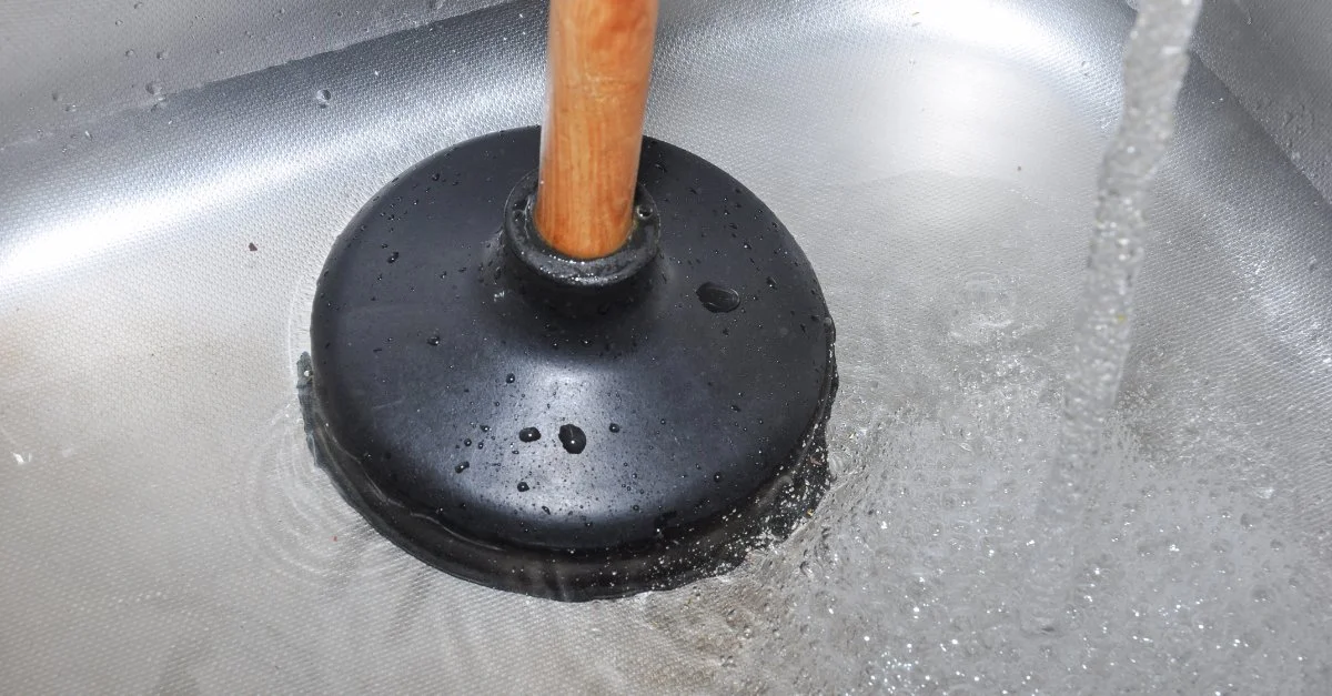 A sink full of water with more water being poured in as a black plunger is used to clear a clogged drain in Vancouver, BC.
