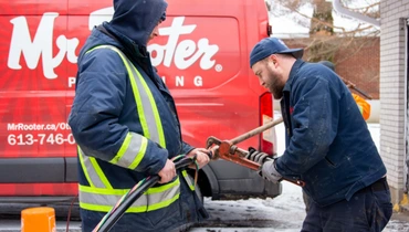 Two professional plumbers operating drain cleaning equipment beside a branded service vehicle in a residential area.