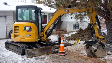 A small excavator digging near a residential property to repair underground plumbing infrastructure.