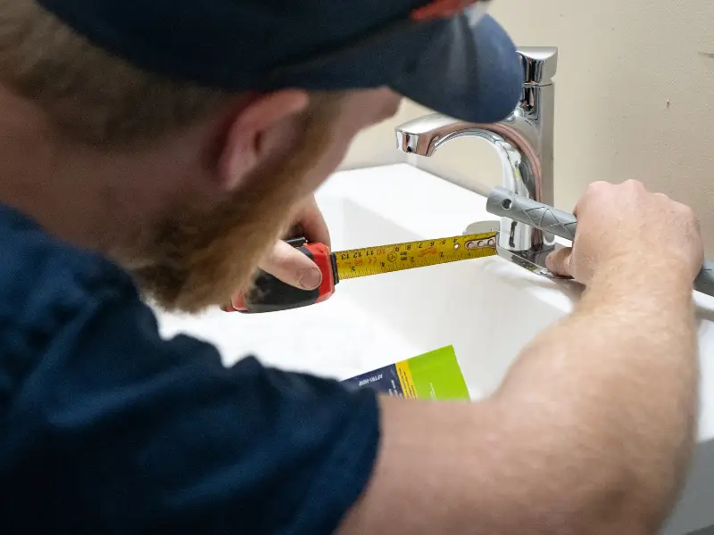 Plumber measuring the distance of a bathroom sink faucet using a tape measure during installation or inspection.