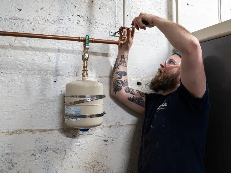 Plumber tightening a copper pipe connection near a thermal expansion tank during a plumbing system inspection.