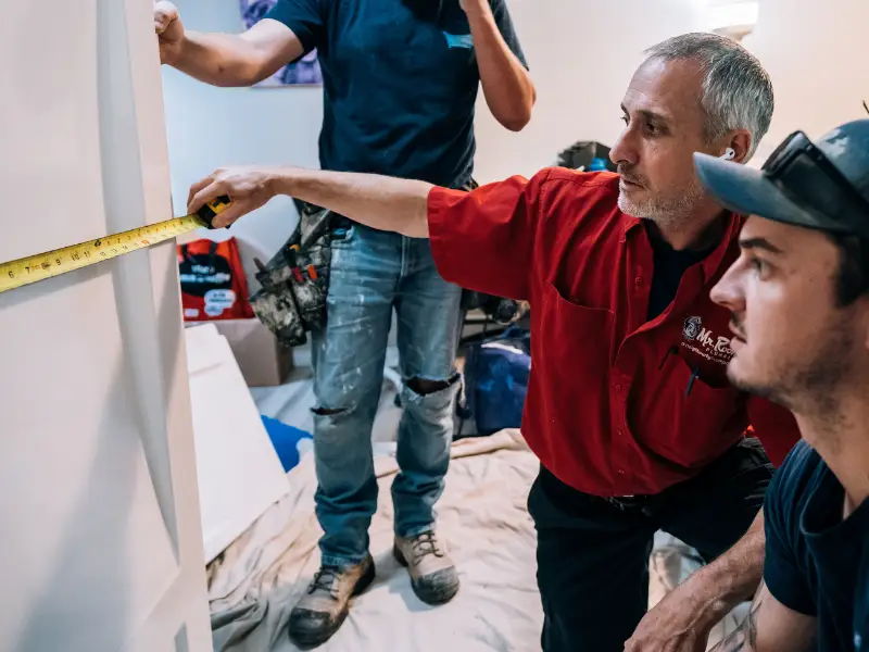 Plumber measuring a wall while discussing plumbing layout or installation details with a teammate.