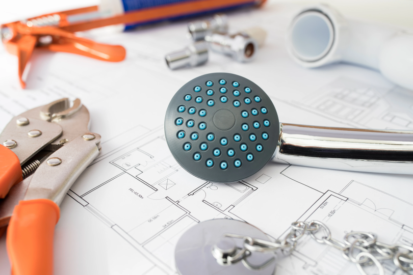 Plumbing tools, shower head, and fittings arranged atop a blueprint of a house symbolizing simple home plumbing jobs for Canadian homeowners.