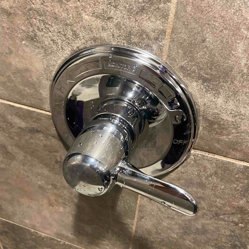 A simple chrome shower handle, installed over tan ceramic tiles, shown in the off position.